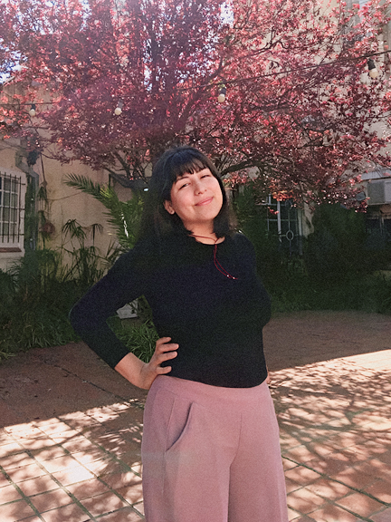 Foto de Fátima sonriendo con remera negra y pantalón rosa, en un patio cerrado, con un arbol rosa de fondo.